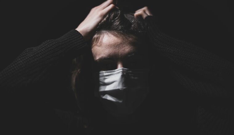 A woman slightly lit in front of the camera wearing a white surgical mask and holding her head